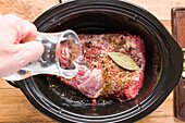 Top view of crop anonymous person adding water on spiced beef brisket with bay leaf and pepper in slow cooker while cooking corned meat