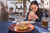 Focused Asian female taking picture of tasty shrimp dish on smartphone while sitting at table with friends during lunch in outdoor cafeteria