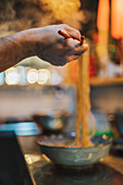 Hand eines gesichtslosen Kochs mit hölzernen Essstäbchen, der eine Nudel über einer Schüssel auf einem unscharfen Hintergrund in einer Ramen-Bar hält