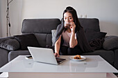 Konzentrierte Asiatin mit Heißgetränk in der Hand schaut auf den Bildschirm eines Netbooks auf dem Tisch mit Gebäck, während sie auf einer bequemen Couch sitzt