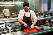 Koch in Schürze beim Schneiden von rohem Fleisch auf einem Brett in der offenen Küche eines Restaurants während der COVID 19-Epidemie