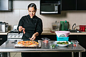 Young ethnic female chef in uniform adding ham on delicious pizza while cooking on kitchen counter in modern cafe