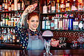 Zufriedene Barkeeperin in Schürze bei der Zubereitung eines Flavor-Blaster-Cocktails am Holztresen einer Bar