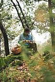 Serious female mycologist sitting on mossy boulder looking at Lactarius deliciosus mushroom in forest with basket