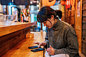 Asiatische Dame in legerem Pullover benutzt ein Handy am Tresen einer traditionellen Ramen-Bar