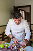 Fokussierter männlicher Koch in Uniform schneidet Fischfilet auf einem Brett, während er am Tisch mit verschiedenen Zutaten in der Küche steht