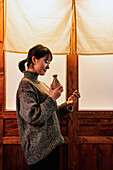Side view of overjoyed Asian woman in sweater standing near door with traditional drink in cafe