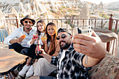 Männliche und weibliche Freunde sitzen mit Cocktails und Bier auf der Terrasse einer Bar und machen ein Selbstporträt mit einer Sofortbildkamera auf der Terrasse in Kappadokien, Türkei