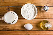 Schüssel mit Mehl und Kanne mit Milch auf Holztisch mit Ei und Öl in heller Küche