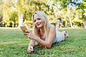 Lächelnde, charmante Frau, die im Park im Gras liegt, ein Selfie mit ihrem Smartphone macht und mit Kopfhörern im Sommer Musik hört