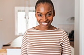Happy young African American female with hair bun looking at camera against table and washing machine in kitchen