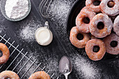 Sweet fried doughnuts served on plate near metal cooling rack and jug of milk on black messy table with powdered sugar