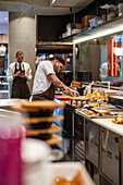 Männlicher Koch bei der Zubereitung von Speisen an der Theke in einer modernen offenen Küche neben einem Kellner, der ein Mobiltelefon im Restaurant während der Arbeit bei COVID Pandemie benutzt