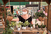 Ältere Einkäuferin mit Textilmaske pflückt Topfpflanzen während der Coronavirus-Pandemie in einem Gartenmarkt