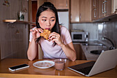 Konzentrierte Asiatin isst ein Pain au Chocolate und surft auf ihrem Netbook, während sie beim Frühstück in einer modernen Küche zu Hause am Tresen sitzt
