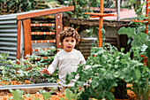 Curly haired boy standing near garden bed with planted sprouts in yard and looking at camera