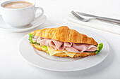 Delicious croissant with ham and cheese placed on white plate near cup of coffee
