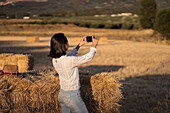Back view of unrecognizable female takin picture on mobile phone while standing in dried field in summer in sunset