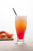 Glass of refreshing Sunrise cocktail with ice cubes and straw served on table with fresh oranges