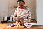 Fröhliche ethnische Frau schneidet frische rote Zwiebeln mit einem Messer auf einem Schneidebrett am Tisch mit einer Schüssel im Haus