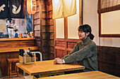 Zufriedene asiatische Frau in legerem Pullover, die an einem Holztisch in einer Ramen-Bar sitzt und mit einem breiten Lächeln in die Ferne schaut