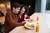Fröhliches multiethnisches Paar isst gesundes Frühstück im Restaurant und surft auf dem Handy