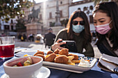 Positive Asian woman with female friend in medical masks watching video on cellphone while sitting at table in outdoor cafeteria