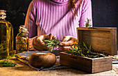 Crop lady cutting rosemary sprigs with scissors at table with fabric and rope near essential oil glass bottles and small chest