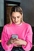 Zufriedene junge Frau in rosa Strickpulli, die in einem hellen Raum auf einem modernen Smartphone surft