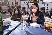 Asian women in protective mask near female friend sitting together at table with dishware in outdoor cafeteria during coronavirus pandemic
