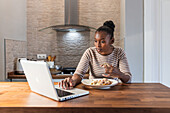 Afroamerikanerin genießt leckeren Patacon mit Topping, während sie mit ihrem Netbook in der Küche zu Hause im Internet surft