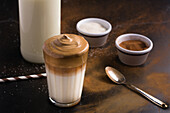 Ein Glas köstlicher Dalgona-Kaffee mit Milch und schaumigem Topping steht auf einem schwarzen, unordentlichen Tisch mit Kakaopulver und Zucker