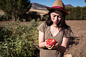 Upset ethnic female farmer in straw hat standing with bitten tomato in field in countryside on sunny day