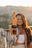Positive junge ethnische Frau lächelt strahlend mit Bierflaschen, während sie eine angenehme Zeit bei Sonnenuntergang auf einer Terrassenbar in Kappadokien, Türkei, genießt
