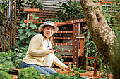 Side view of delighted female gardener in hat sitting near garden bed and spraying sprouts while laughing happily