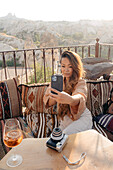 Ethnische Frau auf Kissen sitzend, während sie ein Selbstporträt mit dem Handy am Tisch mit Sofortbildkamera und Cocktail auf einer Terrasse in Kappadokien, Türkei, aufnimmt