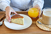 High angle of crop faceless female taking piece of homemade sponge cake from tray with cup of coffee and glass of orange juice during breakfast at home