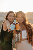 Positive junge multiethnische Freundinnen, die fröhlich lächeln und mit Bierflaschen anstoßen, während sie bei Sonnenuntergang auf der Terrasse einer Bar in Kappadokien, Türkei, eine angenehme Zeit miteinander verbringen