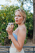 Seitenansicht einer charmanten Frau, die eine kalte, erfrischende Limonade mit Strohhalm trinkt, während sie im Sommergarten chillt und in die Kamera schaut