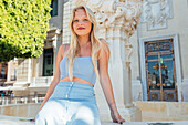 Delighted blonde female in summer outfit sitting in street on sunny day and looking at camera while enjoying summer