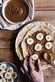 Top view of anonymous person putting pieces of fresh banana on palatable crepes with nut butter on timber table