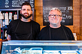 Positive ethnic bearded men in aprons smiling and looking at camera at counter of restaurant with signboards