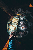 From above hand of crop anonymous person taking glass cup of tea with milk served on tray with flowers in room with sunbeam