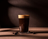 Front view of a black coffee cup with a layer of foam and placed next to some coffee beans on a brown table and wall with dark shadows