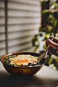 Hand einer anonymen Person, die einen Löffel benutzt, um eine leckere asiatische Suppe mit Nudeln und Spiegelei auf einer Terrasse zu essen