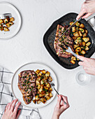 Top view of crop anonymous people eating delicious beef chop with potatoes served on frying pan and plate on white background