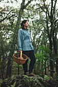 Female with wicker basket seeking wild edible mushrooms while standing on boulders in woods looking away