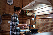 Side view of focused young Asian female in casual clothes cooking yummy pancakes using spatula while standing in kitchen