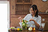 Happy asian woman in casual clothes stirring salad with assorted veggies and fruits with wooden spoon in bowl while cooking in kitchen on sunny day