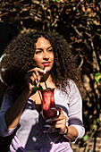 Positive young ethnic female wearing casual outfit enjoying fresh sweet cocktail and looking away while standing in sunny garden